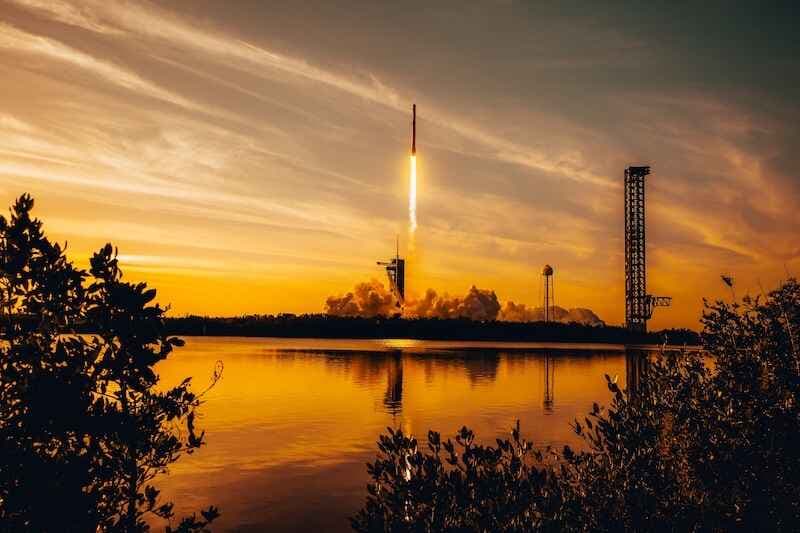 Rocket launch at sunset with fiery trail over water, reflected in golden hues. SpaceX Falcon rocket blasting off. Exploration, technology and space concept.